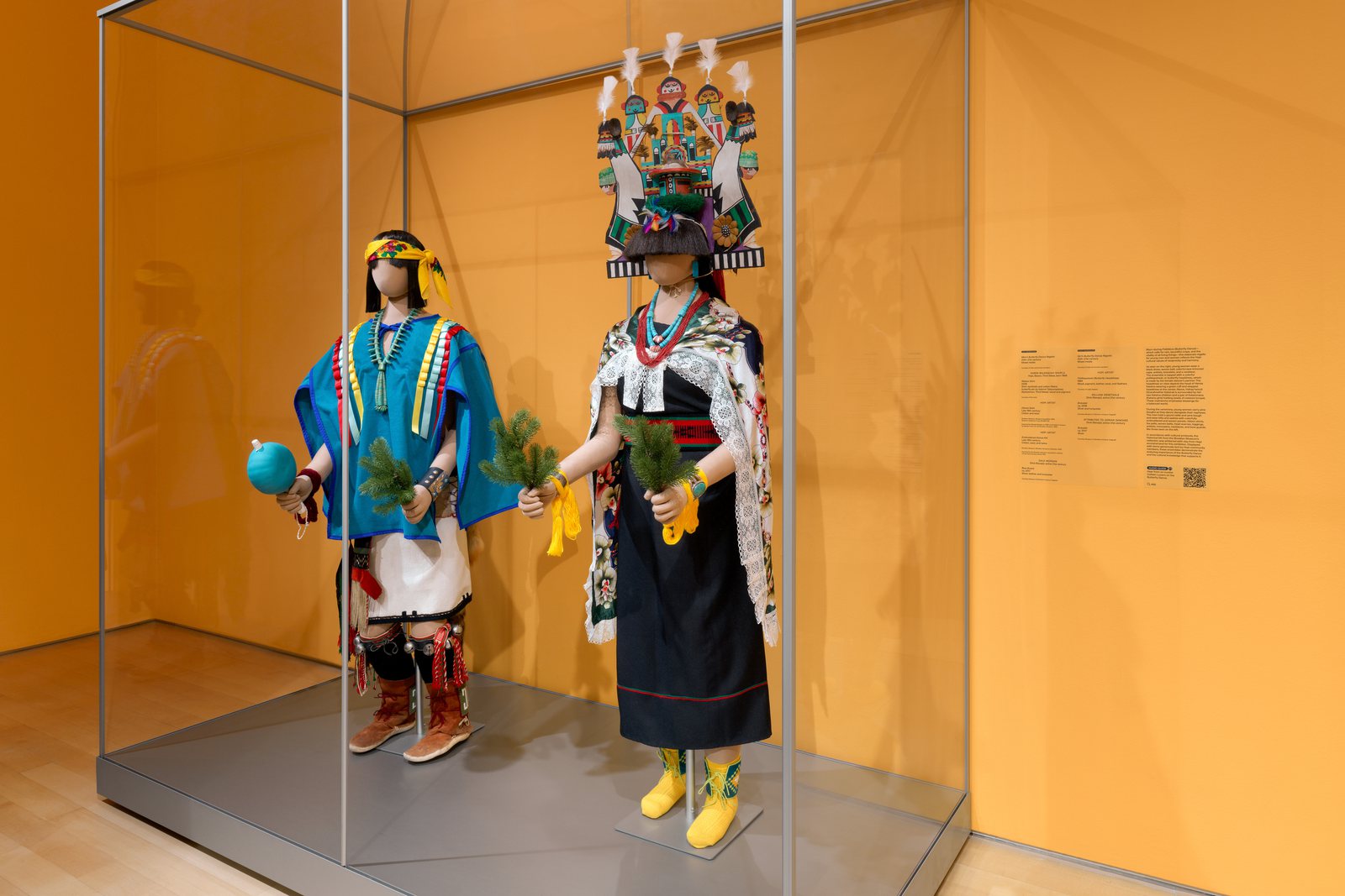 Two Butterfly Dance ensembles, a boy’s and a girl’s with a tall tablita headdress, in a glass case against an orange wall